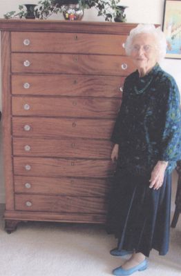 Chest Returns
Beth Mills stands in front of the pine and mahogany chest of drawers made by her grandfather, Charles Johnson, who was an undertaker and cabinet-maker in Mattapoisett at the turn of the 20th century. The chest of drawers remained with her until she passed away in 2008 at the age of 99. Her friends John and Caroline Belloti contacted the Mattapoisett Historical Society after her passing to fulfill her wish that the chest remain with the Mattapoisett Museum, just around the corner from where it was made. 
